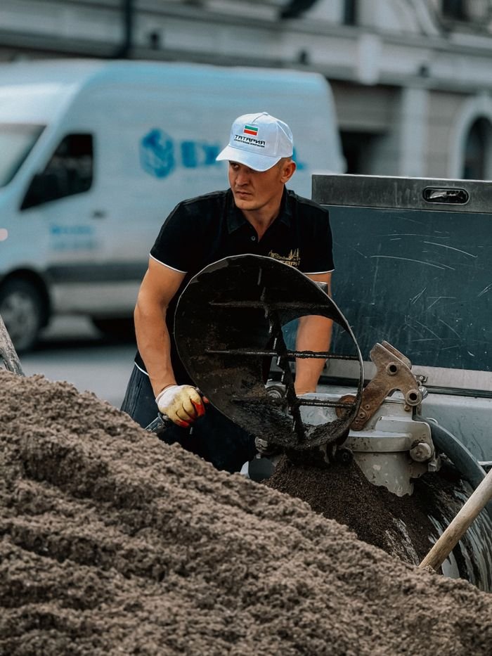 A construction worker operates a sand mixer on a construction site, wearing a branded cap and gloves.